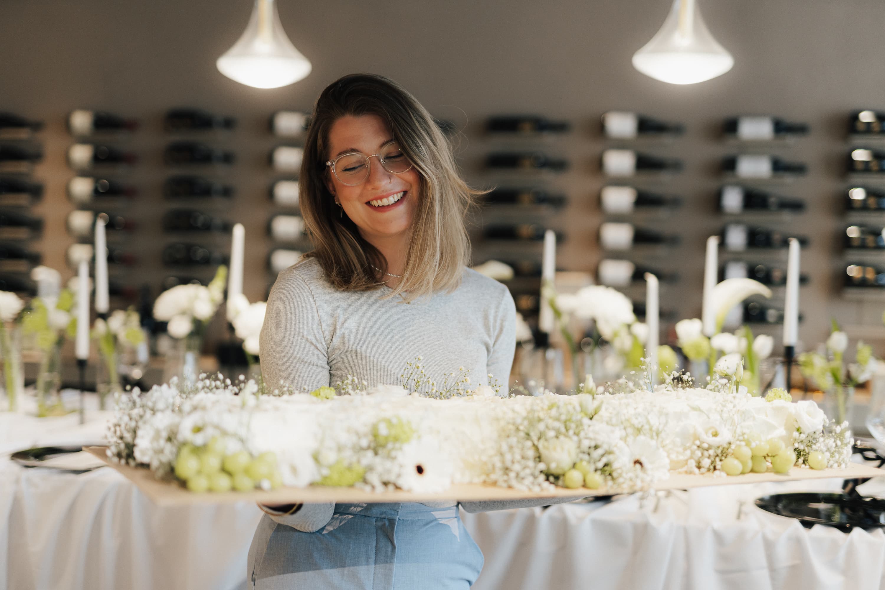 Kerstin Ederer trägt lächelnd ein langes Holzbrett mit einer weißen Sweet-Table-Torte, dekoriert mit Schleierkraut, weißen Blüten und hellen Trauben
